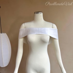 May include: A white off-the-shoulder bridal cape is displayed on a mannequin. The cape is made of a soft, flowing fabric. The text "Pavillon de Veil" is written in cursive in the upper right corner.