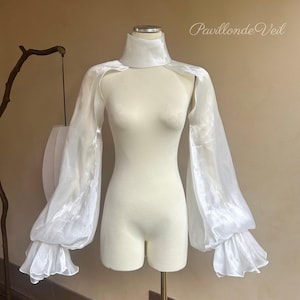May include: A white, sheer, long-sleeved garment with a high collar and ruffled cuffs, displayed on a mannequin. The sleeves are voluminous, and the fabric has a slight sheen. The text "Pavillon de Veil" is visible in the upper right corner.