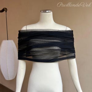 May include: A black, off-the-shoulder garment made of sheer fabric, draped across a white mannequin. The fabric is gathered and layered, creating a textured effect. The background is a neutral tan color, with a hanging lamp and the text "Pavillon de Veil".
