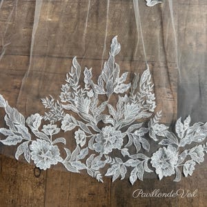 May include: A sheer white bridal veil featuring intricate white floral embroidery and delicate silver sequins. The lace applique displays a detailed botanical pattern of leaves and blossoms along the edge, with the text PavillondeVeil visible.