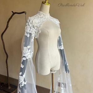 Può includere: Una mantella da sposa bianca con dettagli in pizzo floreale. La mantella presenta maniche lunghe e trasparenti e un collo alto. Il testo "Pavillon de Veil" è visibile in alto a destra. La mantella è esposta su un manichino.