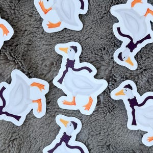 May include: A collection of die-cut stickers featuring a white goose with orange feet and a beak, wearing a dark purple scarf and a matching dark purple beanie. Each sticker depicts the same goose character in a repeating pattern on a grey surface.