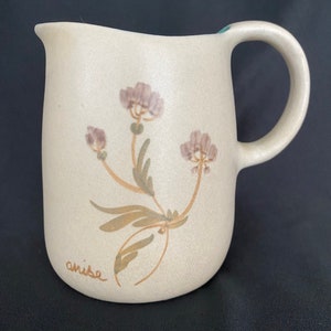 May include: A beige ceramic pitcher with a brown handle and a floral design. The pitcher has a light brown glaze and a light blue rim. The floral design features brown stems and leaves with purple flowers. The word "anise" is written on the bottom of the pitcher.