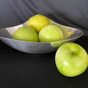 May include: Four green apples in a silver bowl. The bowl is shaped like a crescent moon and has a smooth, reflective surface.