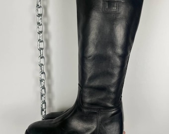 May include: A tall, black leather boot with a rounded toe and a high shaft. The boot features a rectangular detail near the top and is held up by a hand. The boot's height is approximately 16 inches.