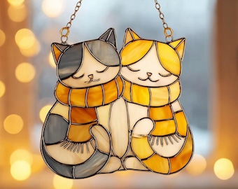 Puede incluir: Atrapasoles de gato de vidriera con dos gatos con los ojos cerrados, que llevan bufandas amarillas y naranjas. Un gato es gris y blanco, el otro es amarillo y blanco. Los gatos están conectados por una cadena dorada para colgar.