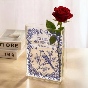 May include: A clear acrylic vase with a blue floral and bird design, featuring the text "TO KILL A MOCKINGBIRD by Harper Lee." A red rose with green leaves is placed inside the vase. Two books are visible in the background.
