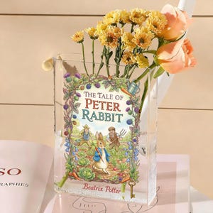May include: A clear glass vase featuring the cover art from "The Tale of Peter Rabbit" by Beatrix Potter. The vase holds yellow chrysanthemums and peach roses. The book cover art depicts Peter Rabbit in a garden scene.