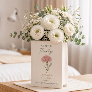 以下が含まれることがあります： 白いセラミック製の本型の花瓶に、白い花と緑の葉のブーケが入っています。花瓶には「CHAPTER Thirty」と「JANUARY Carnation」の文字と花のイラストが描かれています。「Every chapter brings new growth」というフレーズも印刷されています。
