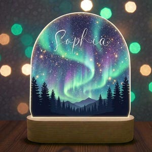 Puede incluir: Una luz de noche decorativa que presenta una vívida representación de la aurora boreal, con luces verdes y púrpuras arremolinadas sobre un bosque y montañas silueteadas. El nombre "Sophia" está escrito en cursiva. La luz se encuentra sobre una base de madera.