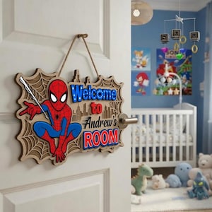 Peut inclure: Une plaque de porte en bois avec un motif de toile d'araignée en relief et une illustration colorée d'un super-héros rouge et bleu. La plaque porte l'inscription Welcome to Andrew's ROOM en lettres bleues et rouges, suspendue par une cordelette.