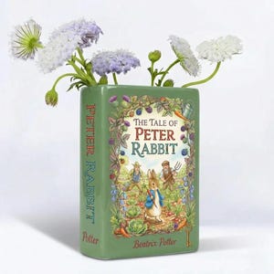 Puede incluir: Un jarrón de cerámica verde con forma de libro, con la portada de "The Tale of Peter Rabbit" de Beatrix Potter. El jarrón contiene flores blancas y lilas. El lomo del libro dice "Peter Rabbit".