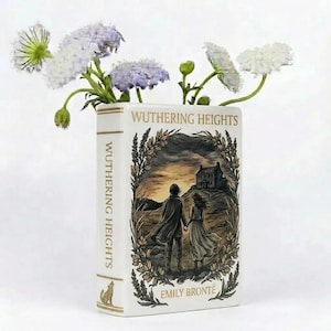 Può includere: Un vaso in ceramica bianca a forma di libro, intitolato "Wuthering Heights" di Emily Brontë. La copertina raffigura una coppia che cammina verso una casa. Il vaso contiene fiori bianchi e viola.