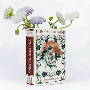 Op de afbeelding: Een witte keramische vaas in de vorm van een boek, met de titel "Gone With The Wind" en een illustratie van een stel. De vaas heeft een bordeauxrode rug met de titel en de naam van de auteur. Witte en paarse bloemen zijn in de vaas gerangschikt.