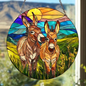 Op de afbeelding: Rond gebrandschilderd glaspaneel met twee ezels in een veld met een zonsondergang op de achtergrond. Het kunstwerk gebruikt tinten bruin, oranje, geel, groen en blauw. Een metalen ketting is bevestigd om op te hangen. Decoratief paneel.
