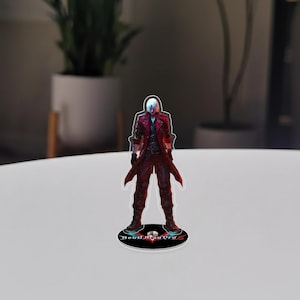 May include: A clear acrylic figure of a character from Devil May Cry 5, standing on a black base. The character is depicted in a long red coat, black trousers, and white hair. The base has the game's title in a stylized font.