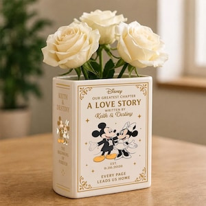 Può includere: Un vaso in ceramica color crema a forma di libro con scritte dorate e illustrazioni di Topolino e Minnie. La parte anteriore riporta la scritta A LOVE STORY WRITTEN BY Keith & Destiny, mentre il dorso indica KEITH & DESTINY. Tre rose bianche.