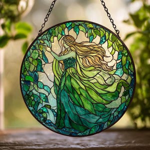 May include: Round stained glass panel featuring a woman in a flowing green dress, surrounded by green foliage. The woman has long blonde hair and is reaching towards a branch. The panel is framed in black and suspended by a chain.