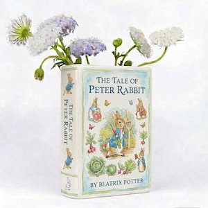 Puede incluir: Un jarrón de cerámica blanco con forma de libro, con ilustraciones y texto de "The Tale of Peter Rabbit" de Beatrix Potter. El jarrón contiene flores blancas y moradas.