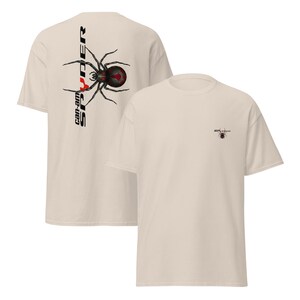 Pode incluir: Duas camisetas bege. Uma apresenta um gr&aacute;fico de aranha preta e vermelha com o texto "can-am SPYDER" verticalmente no lado esquerdo. A outra camisa tem um gr&aacute;fico de aranha menor.