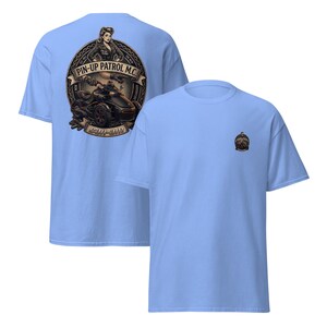Puede incluir: Camisetas de color azul claro con un dise&ntilde;o gr&aacute;fico. Una camiseta presenta un gran emblema circular con una pin-up, una motocicleta y el texto "PIN-UP PATROL M.C." y "CAN-AM". La otra camiseta tiene un emblema m&aacute;s peque&ntilde;o.