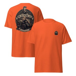 Puede incluir: Camiseta naranja con un gr&aacute;fico en la espalda que representa una pin-up, una motocicleta y el texto "PIN-UP PATROL M.C." Un gr&aacute;fico m&aacute;s peque&ntilde;o est&aacute; en la parte delantera. La camiseta est&aacute; hecha de un material suave.