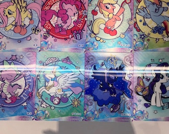 May include: A collection of colorful trading cards featuring cartoon ponies in various poses. Each card has a circular design with a different pony, surrounded by decorative elements and text. The cards have a holographic background.