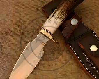 May include: A hunting knife with a polished steel blade and a handle made of bone and antler. The knife is accompanied by a brown leather sheath with a snap closure. The blade is approximately 8 inches long.