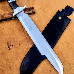 Puede incluir: Un cuchillo grande con una hoja plateada, un mango negro y una funda de cuero negro. La hoja tiene texto grabado. El cuchillo está sobre una superficie de madera.