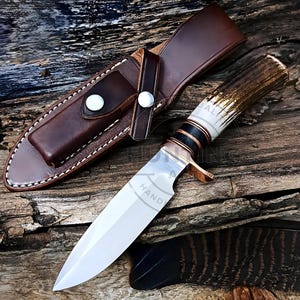 Op de afbeelding: Een handgemaakt jachtmes met een gepolijst stalen lemmet en een handvat van geweien met donkere accenten. Het mes ligt naast een bruine leren schede met witte stiksels en een drukknoopsluiting. Op het lemmet staat het woord "HAND".