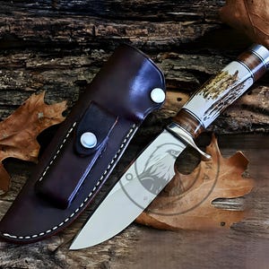 Op de afbeelding: Een jachtmes met een gepolijst stalen lemmet en een handvat met een combinatie van licht- en donkerbruine materialen. Het mes wordt geleverd met een donkerbruine leren schede met witte stiksels en een drukknoopsluiting. Het lemmet heeft een geëtst ontwerp.