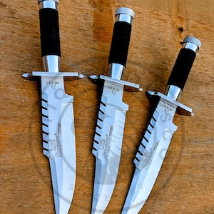 May include: Three survival knives with silver blades and black handles are displayed. The blades have a serrated edge and the text "LIFE Support System I". The handles are wrapped in a textured black material. The knives are on a wooden surface.