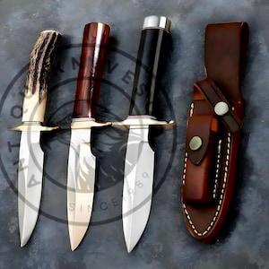 Op de afbeelding: Drie jachtmessen met verschillende handvatontwerpen: gewei, donker hout en zwart. Elk mes heeft een zilveren lemmet en een messing bolster. Een bruine leren schede met een drukknoopsluiting is ook aanwezig.