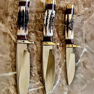 Op de afbeelding: Drie jachtmessen met goudkleurige lemmeten en handvatten. De handvatten hebben een combinatie van donkerbruine, crèmekleurige en gouden accenten. De messen zijn van groot naar klein gerangschikt en tonen hun verschillende maten en ontwerpen.