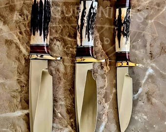 May include: Three hunting knives with gold-colored blades and handles. The handles feature a combination of dark brown, cream, and gold accents. The knives are arranged from largest to smallest, showcasing their varying sizes and designs.