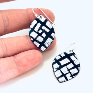 May include: A pair of leaf-shaped earrings with a black and white geometric pattern. The earrings have silver-colored hooks. The design features a grid-like pattern of white shapes on a black background. The earrings are displayed against a white background.