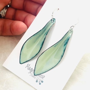 May include: A pair of dangle earrings with a light green enamel finish and a silver hook. The earrings are shaped like a teardrop with a dark green outline.