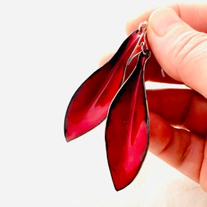 May include: A pair of red enamel earrings shaped like leaves. The earrings have a silver hook closure.