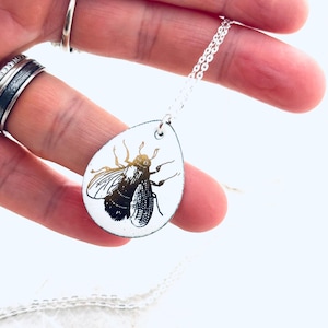 May include: A silver chain necklace with a teardrop-shaped pendant featuring a black and white illustration of a bee.