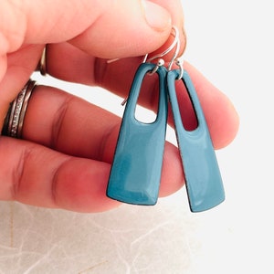 May include: A pair of blue enamel earrings with a rectangular shape and a cutout in the center. The earrings are suspended from silver hooks.