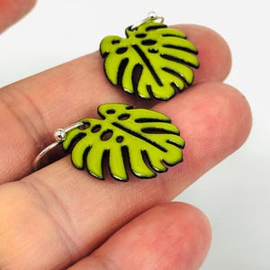 May include: A pair of lime green leaf-shaped earrings with black outlines. The earrings are made of a shiny material and have a silver hook. The earrings are a tropical, botanical theme.