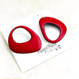 May include: A pair of red enamel earrings with a unique, asymmetrical, oval shape. The earrings have a black outline and a silver hook.