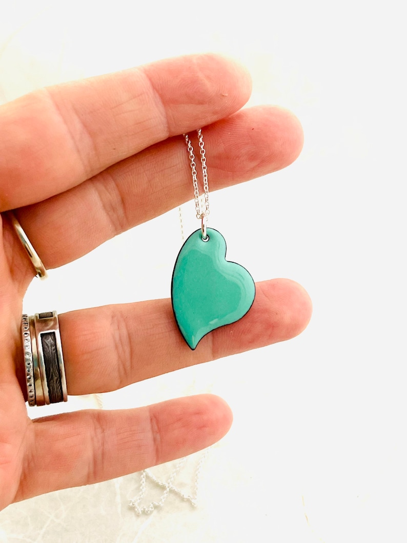 May include: A silver chain necklace with a turquoise blue heart pendant. The heart is slightly asymmetrical and has a black outline.