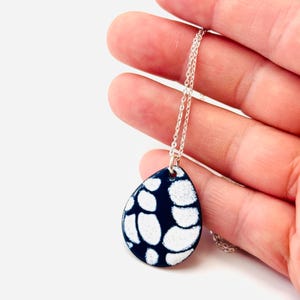 May include: A teardrop-shaped pendant necklace with a silver chain. The pendant is navy blue with a white, organic pattern. The necklace is held in a hand against a white background.