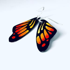 May include: A pair of drop earrings shaped like butterfly wings. The wings feature a black base with vibrant orange and yellow gradient patterns, mimicking the appearance of monarch butterfly wings. Each earring has a silver-toned metal hook.