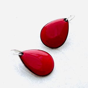 May include: A pair of teardrop-shaped earrings featuring a glossy, vibrant red enamel finish on the front surface. Each earring is attached to a simple, thin silver-toned metal hook wire, providing a clean and minimalist look for these accessories.