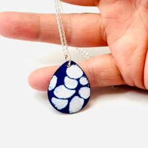May include: A silver necklace featuring a teardrop pendant with a dark blue enamel base and a white, organic pattern. The pendant is suspended from a delicate silver chain, creating a striking contrast of colors and textures.