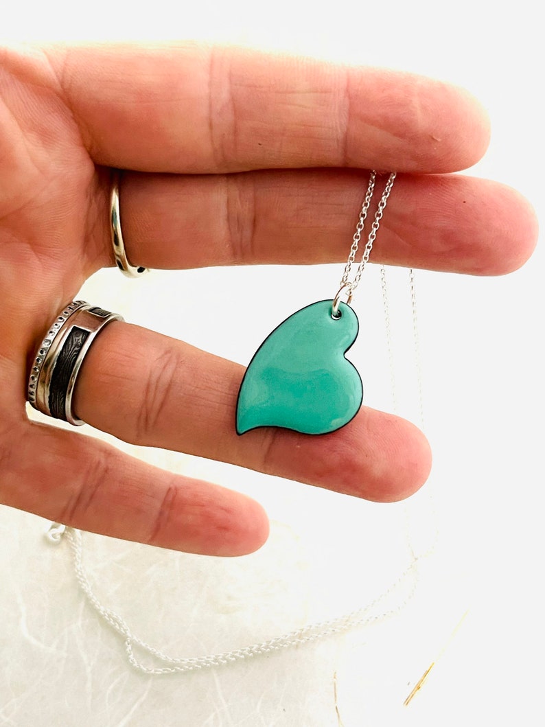 May include: A silver chain necklace with a teal blue heart pendant. The heart is slightly asymmetrical and has a black outline.