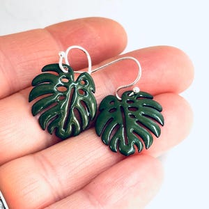 May include: Dark green leaf-shaped earrings with silver hooks. The earrings are a glossy, enamel-like finish, and the leaf design has intricate details. The earrings are held up by a person's hand.