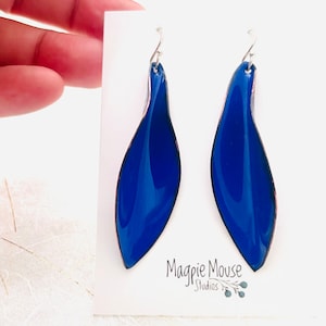 May include: A pair of blue enamel earrings with silver hooks. The earrings are shaped like a leaf and have a glossy finish. The earrings are displayed on a white card with the text "Magpie Mouse Studios".